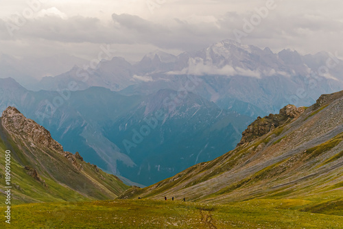 Photo from the back of hikers walking on green grasslands in the Tianshan Mountains in Xinjiang, China. There is snow on the peak with copy space on top. Idea for adventure activity wallpaper.