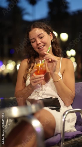 Young woman in summer dress with cocktail in her hands sitting in square on hot evening, she came on vacation to Spain and goes to parties. Nightlife and bars in Barcelona. High quality 4k footage