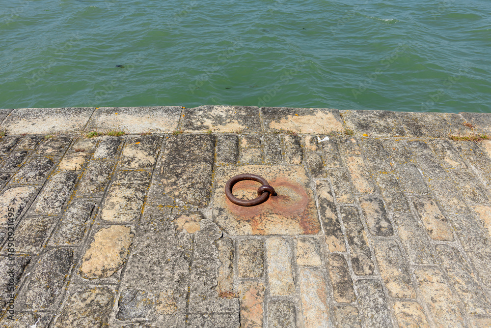Fototapeta premium A weathered iron mooring ring is set into an aged stone quay by green harbor water in Locmariaquer. Sunlit masonry displays rough textures and subtle earth tones, evoking a maritime atmosphere.