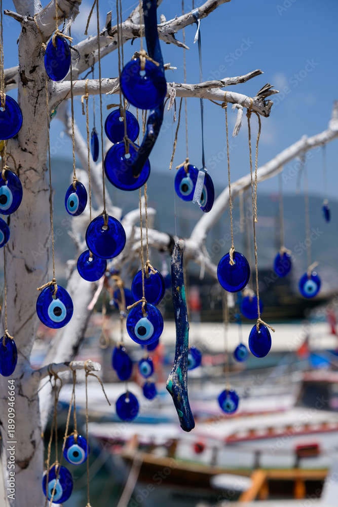 Fototapeta premium Decorative Nazar amulets, talismans hanging on tree in seaport, handmade beads symbolizing traditions, protection from evil. Blue eye-shaped pendants hanging on weathered piece of driftwood above pier