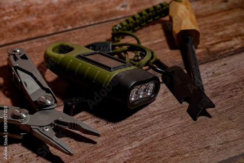 Survival gear kit, including a multitool, a solar flashlight, and a fire starter, arranged on a rustic wooden surface