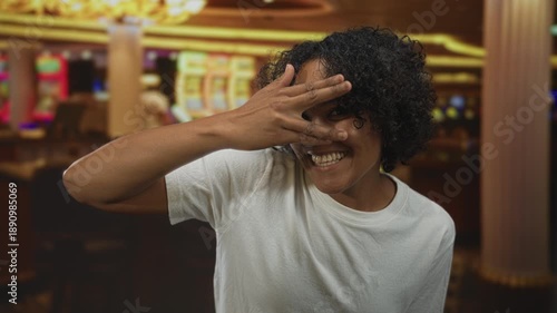 Woman covering eyes with hand and smiling behind fingers in casino building beside slot machines; excitement.