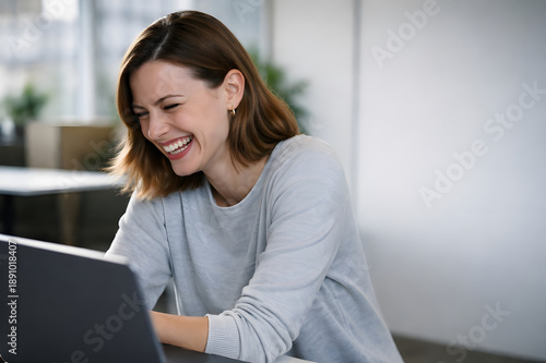 Candid shot of a happy young woman laughing sincerely while looking at her laptop screen in a modern office or home workspace.