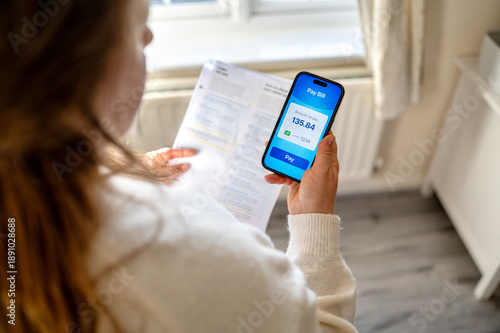 Woman checking utility bills on smartphone while holding paper invoice at home. Household expenses and budgeting concept.