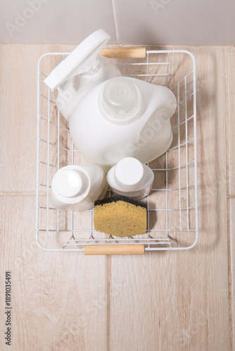 Cleaning products in white bottles in a white basket on a tiled floor. White bottles of detergent for washing and cleaning in a wire basket on the white
