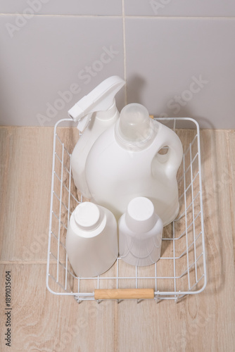 Cleaning products in white bottles in a white basket on a tiled floor. White bottles of detergent for washing and cleaning in a wire basket on the white