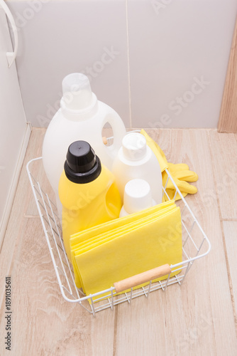 Bottles of detergents for washing and cleaning with gloves, sponges, and rags in a wire basket on the floor