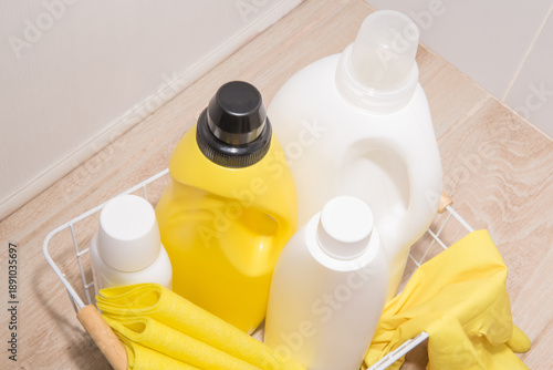 Bottles of detergents for washing and cleaning with gloves, sponges, and rags in a wire basket on the floor