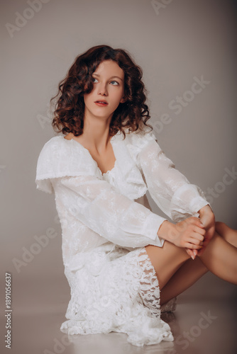 The model is a young beautiful brunette girl with curls on a gray background in a white blouse and lace skirt