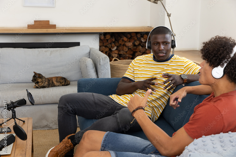 Fototapeta premium Diverse male friends recording podcast on sofa in living room with microphones and headphones
