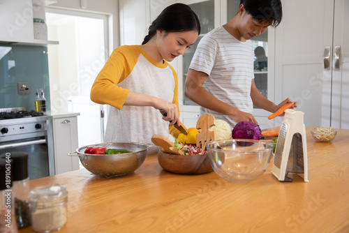 Asian couple slicing bell pepper into salad bowl and peeling carrots on home kitchen island