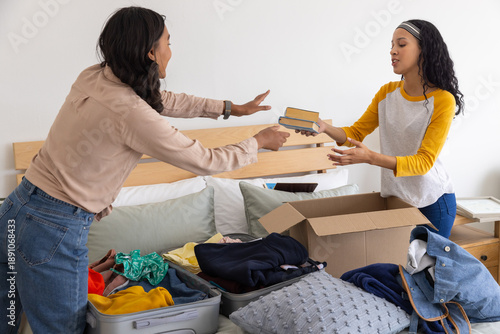 Diverse mom and teenage daughter passing hardcover books across bed in bedroom with suitcase