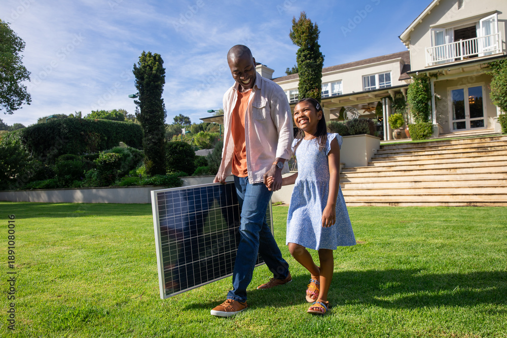 Obraz premium Diverse father and daughter walking across lush lawn at suburban home, carrying solar panel