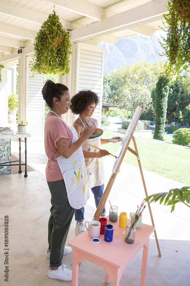 Obraz premium Diverse mother and daughter painting on wooden easel on veranda in aprons amid hanging plants