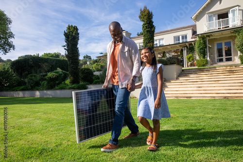 Diverse father and daughter walking across lush lawn at suburban home, carrying solar panel