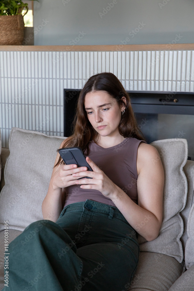 Obraz premium Female in her twenties holding smartphone and leaning on beige sofa by fireplace in living room