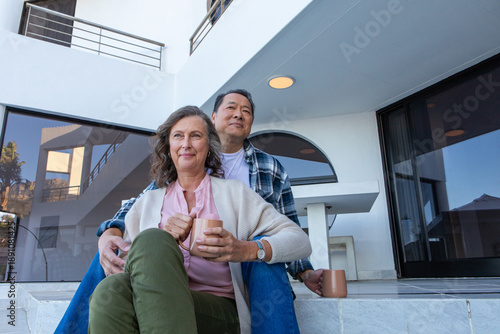 Φωτογραφία Senior couple sitting on steps under balcony at home holding mugs and gazing tho
