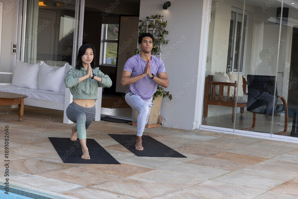 Fototapeta premium Diverse yoga partners holding forward lunge on mats at poolside terrace beside sliding doors