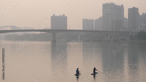 Hazy urban river scene with a bridge and high-rise buildings in the background, two people paddleboarding on calm water at dusk in Nanning, China. Atmospheric cityscape and relaxing vibe