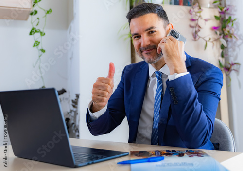 Tableau sur toile Young entrepreneur looking very pleased, nodding his head while talking on the phone and working on his laptop