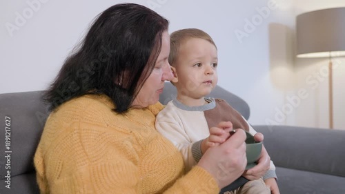 Grandmother feeding grandson with a spoon on the sofa
