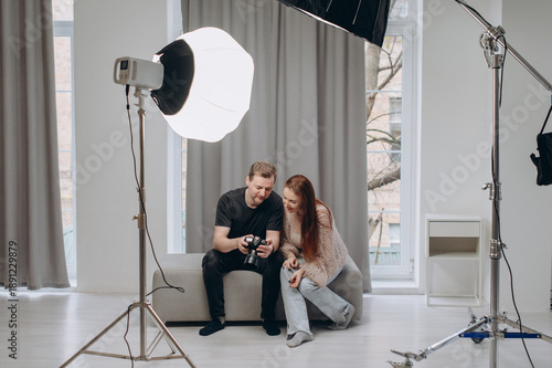 A photographer and a female model review photos together on a camera screen during a studio photoshoot with professional lighting.