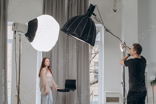 A photographer adjusts the lighting in the studio to take portraits of a young redhead model.