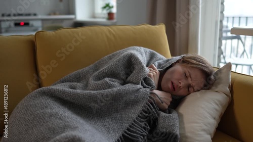 Sad lost young woman feels unwell lying on sofa wrapped in wool plaid, showing first signs of cold and flu symptoms. Sick female suffering from chills, curling on couch with fever by virus infection