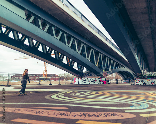 Urban skateboarding scene under a modern arched bridge, featuring colorful circular ground art, skaters in motion and an industrial city backdrop on an overcast day