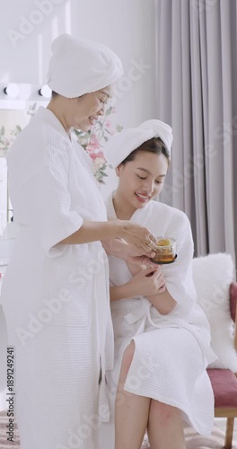 Asian senior mother preparing honey facial mask with daughter in white robes during skincare routine, expressing care, bonding, and natural beauty in warm spa environment