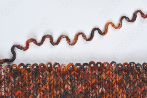 A horizontal flat lay shows a dark, multicolored, hand-knitted wool swatch with a decorative rolled edge below a long, wavy strand of matching yarn on a plain gray background