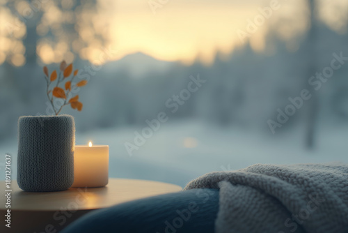 A cozy winter scene with a lit candle, knitted pot, and blanket near a window overlooking a snowy landscape