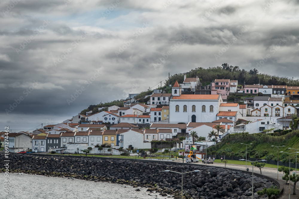 Fototapeta premium Panoramic view of a Ribeira Quente village with white houses and orange tiled roofs, centered around a local church. The foreground features a volcanic stone seawall along the Atlantic coast.