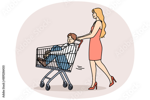 Woman carries man in supermarket carts, for concept of buying boyfriend and financially motivated marriages. Girl gives capricious boyfriend ride on buyer cart because guy doesnt want to go shopping