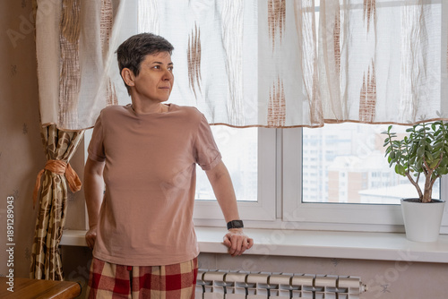 Tablou pe pânză Middle-aged woman leans on a windowsill and looks outside through sheer curtains in a modest apartment, soft daylight interior scene conveying thoughtfulness, home isolation and quiet daytime pause