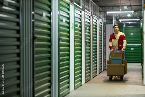 Fotografia Young adult Caucasian man pushing cart loaded with cardboard boxes through corri