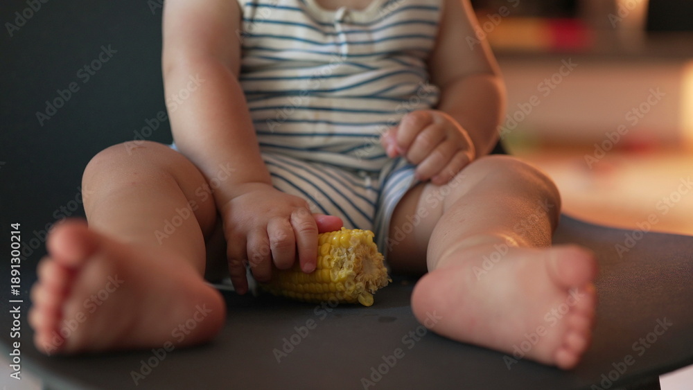 Obraz premium Toddler sits with legs outstretched, holding corn cob while seated on black chair, showcasing relaxed grip and foot positioning in casual indoor space