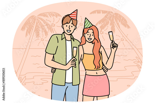 Honeymoon happy couple drinking champagne, standing on beach of sunny resort, wearing birthday caps. Romantic guy and girl celebrate beginning of honeymoon on ocean shore with palm trees