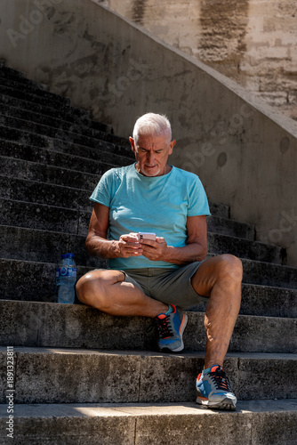 Senior man sitting on staircase checking smartphone after outdoor exercise, staying hydrated