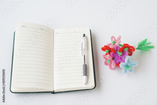 Open blank notebook with pen placed on white background next to colorful handmade craft flowers. Creative workspace concept for planning, writing, education, DIY art, imagination and inspiration 