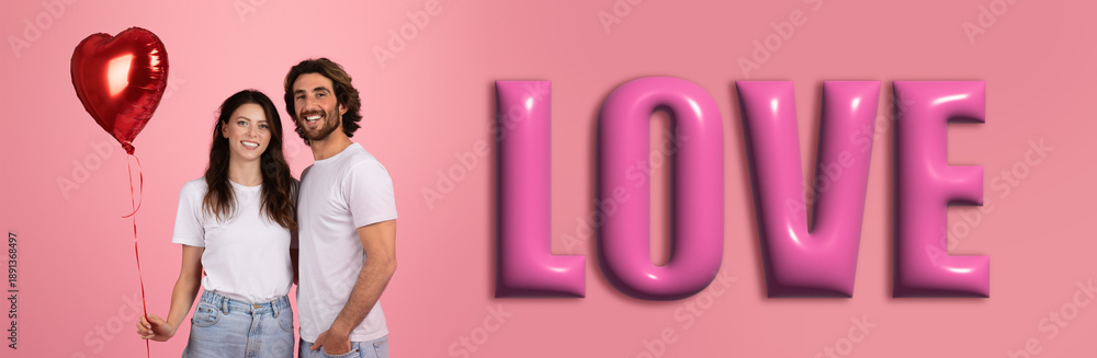 Fototapeta premium A couple stands together holding a red heart-shaped balloon. They pose next to the large word love on a pink background. The scene shows happiness and connection between them.