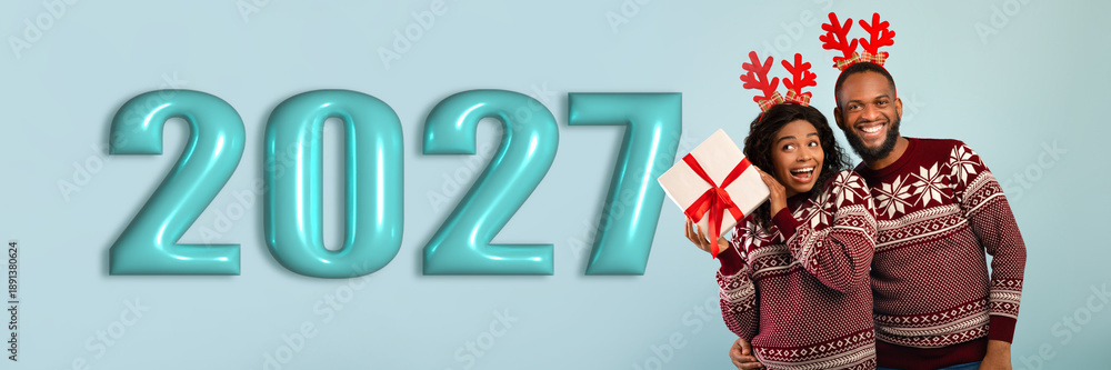 Fototapeta premium A couple stands together, smiling and holding a gift. They wear matching holiday sweaters and have festive reindeer antlers. The backdrop shows the year 2027 in large numbers.