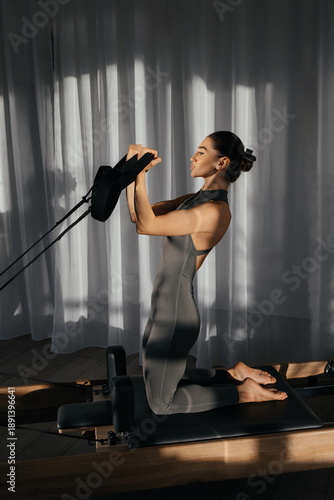 Pilates reformer training machine. Fit slim woman trains on Pilates reformer in sunlit studio. Hands gripping straps and controlled movements highlight full body engagement 