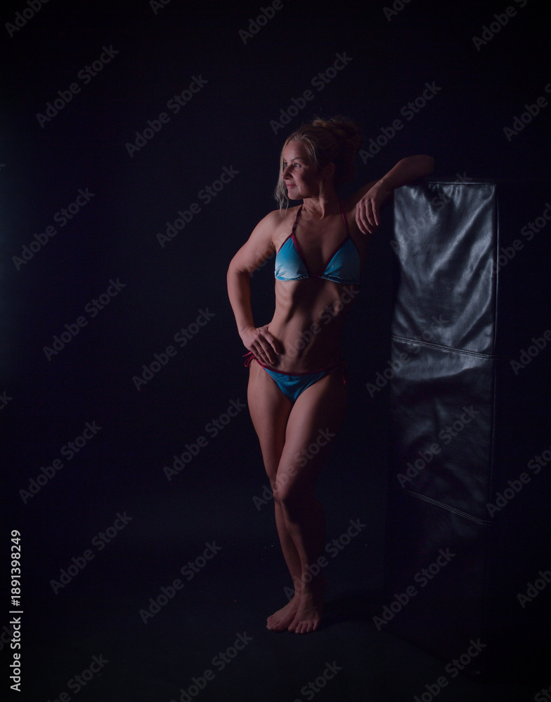 Fototapeta premium Muskulöse, trainierte Bodybuilderin im Best-Age-Alter posiert selbstbewusst im Studio. Symbol für Fitness, Kraft, Disziplin und aktives Älterwerden. Dunkler Hintergrund, Fokus auf Körperform und Stärk