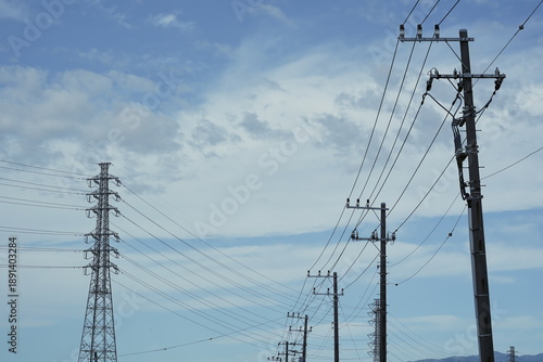 Obraz na płótnie 青空の下に並ぶ電柱と遠くに見える大きな送電鉄塔の風景 幾重にも重なる電線が空を横切る日本の社会インフラを感じさせる日常の景色