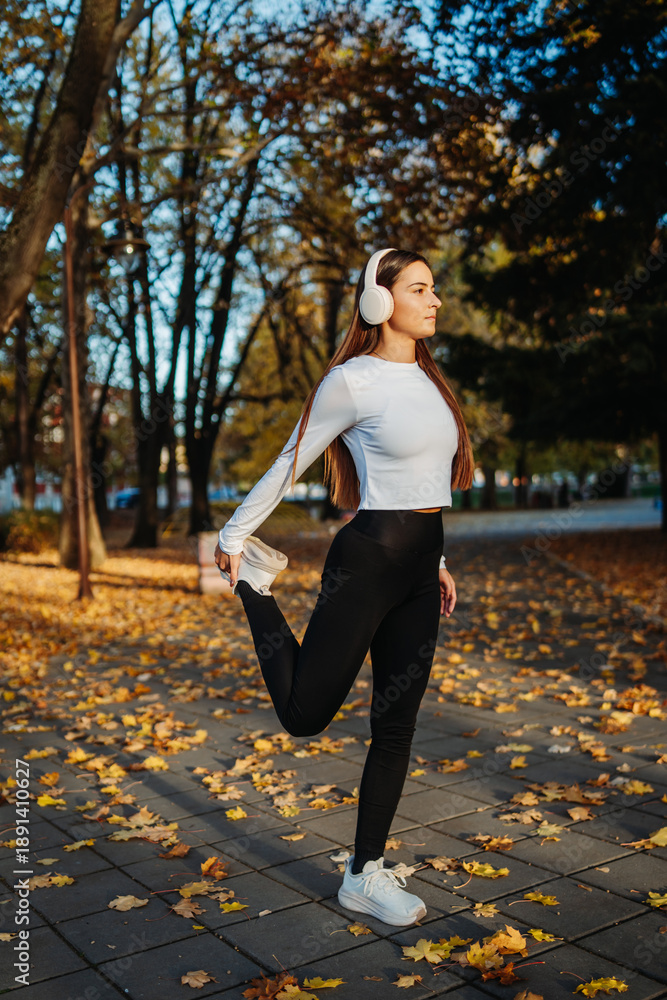 Fototapeta premium Woman stretching in autumn park wearing wireless headphones
