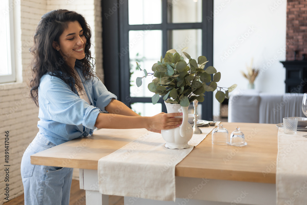 Naklejka premium Modern home interior design, Scandinavian style concept. Beautiful young brunette woman arranging plants in vase, decorating dining room table, creating cozy atmosphere in her apartment, copy space