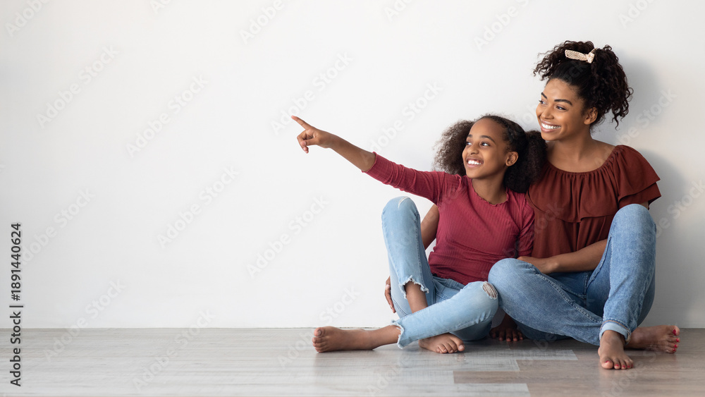 Fototapeta premium Cheerful african american young woman mother and school girl daughter in similar outfits sitting on floor at home, hugging, smiling, teen girl pointing at copy space for advertisement, panorama