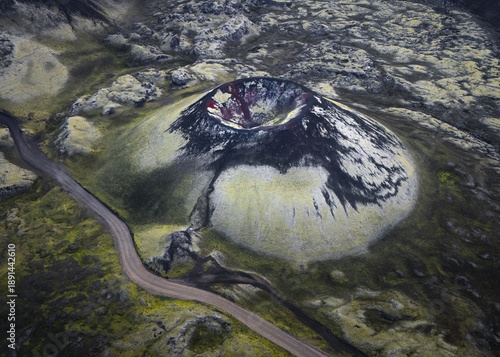 Aerial view of the stark, volcanic landscape, where a dark, imposing crater dominates the scene, surrounded by patches of green moss and a winding road, Myvatn, Thingeyjarsveit, Iceland.