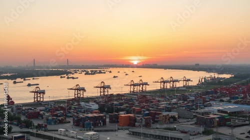 A port with cranes and ships sits beside a river as the sun sets over Wuhan, China. Sunrise over a busy shipping port in Wuhan, China, with cargo cranes and ships on the river